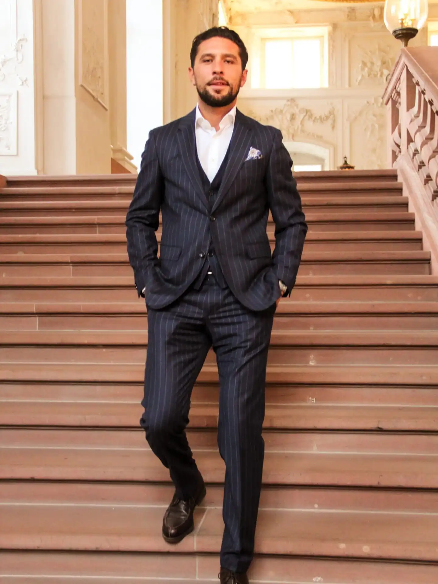 Man in a dark pinstripe suit walking down stone stairs in an ornate building.