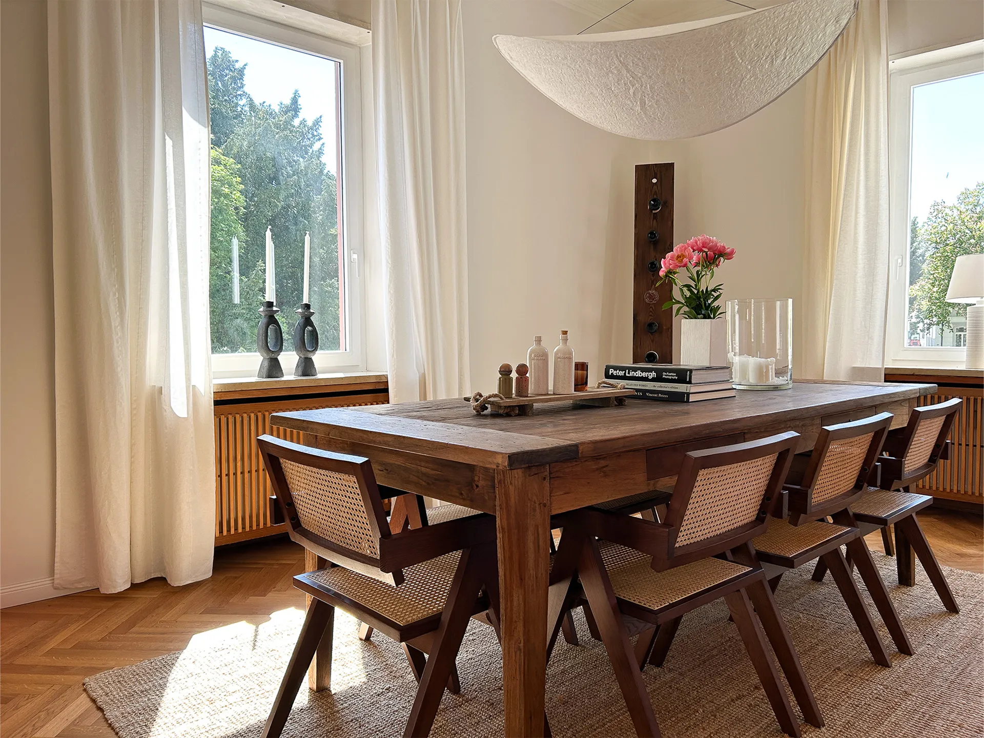 Sunlit dining room with a wooden table surrounded by six chairs, decorated with books, pink flowers, and candles.