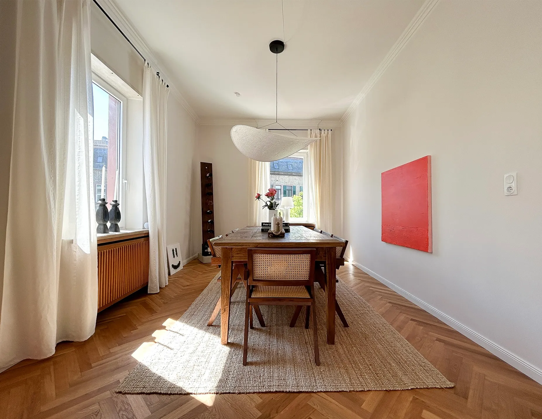 Bright dining room with wooden table and chairs on a beige rug, white curtains framing windows, pink flowers in a vase, and a red painting on the wall.